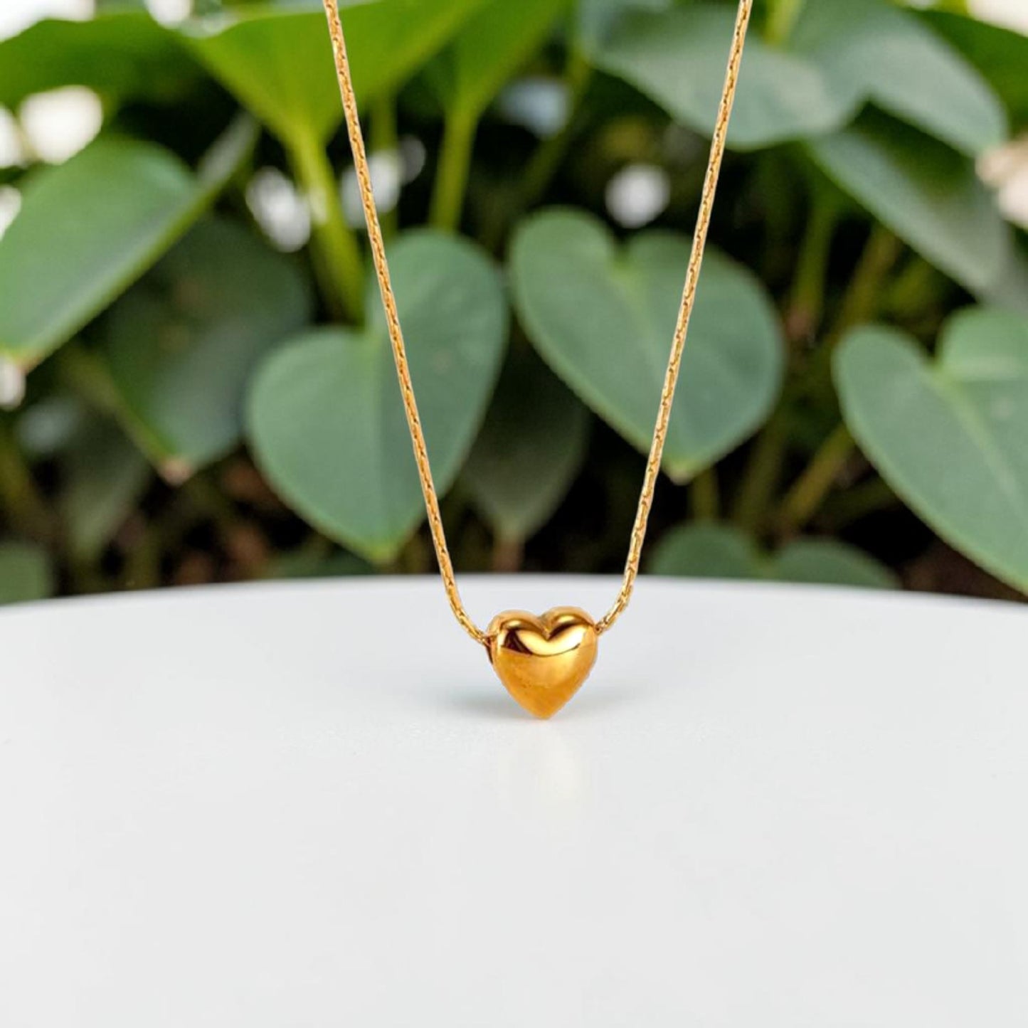 Gold heart-shaped necklace on a white surface with green leaves in the background. 

18K gold plated stainless steel heart pendant necklace - tarnish free, hypoallergenic, water resistant